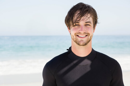 Man standing near waterline on sandy beach wearing black wetsuit and smiling, copy space