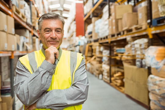 Senior male standing in warehouse aisle inspecting inventory on racks wearing safety vest