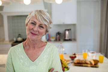 Senior woman standing at kitchen island preparing breakfast with orange juice, toaster and fruit