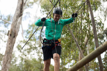 Man balancing on suspended wooden beam in forest, gripping steel cables with helmet and harness
