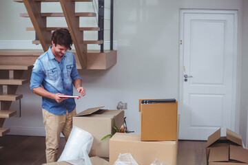 man unpacking cardboard boxes beside wooden staircase checking red-framed tablet on hardwood floor