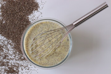 Chia custard in a glass bowl with whisk, surrounded by chia seeds on a white surface