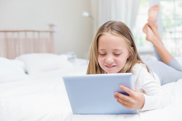 Schoolgirl lying on bed holding tablet under sheer curtains beside lamp in bedroom