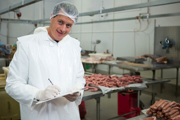 Male inspector wearing coat examining sausage trays at meat plant holding clipboard, copy space