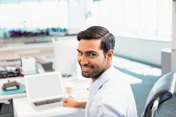 Asian man turning in swivel chair smiling at laptop on white desk in open-plan office