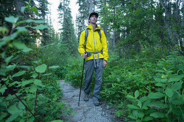 Male hiker standing on rocky forest trail in yellow rain jacket with backpack, boots, trekking pole