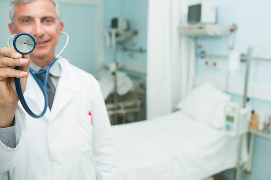 Senior doctor offering stethoscope to viewer in hospital room with bed, monitor, copy space - Powered by Adobe