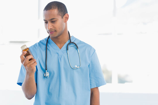African American male doctor in light blue scrubs and stethoscope examining pill bottle in clinic