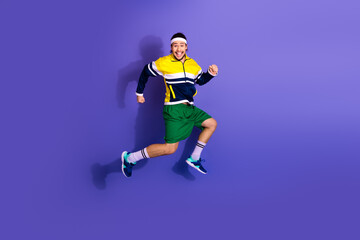 Cheerful male athlete dressed in retro sportswear jumping mid-air on purple background expressing energy and joy