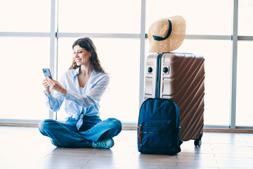 Female traveler sitting at airport terminal with phone and luggage, capturing moments of her...