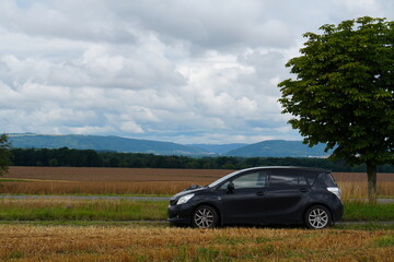 Dark Hatchback Car Parked by a Harvested Field with Rolling Hills Under a Cloudy Sky
