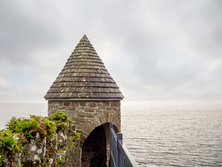 Fototapeta premium A small stone building with a pointed roof sits on a pier overlooking the ocean. The sky is overcast, and the water is calm. The building appears to be a small shelter or castle lookout point