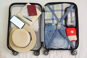 top view of open suitcase on bed for packing, preparing for summer holidays abroad trip with passport, map, hat, travel medicine first aid kit, clothing, phone mockup with copy space for advertise