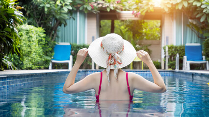 back view of woman in pink swimsuit with straw hat relaxing in swimming pool at hotel, enjoy summer holiday and vacation at tropical country, sunbathing wellness lifestyle concept.