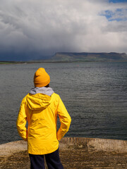 Girl tourist in yellow hat and jacket is standing on a pier and looking at ocean and Benbulbin flat...