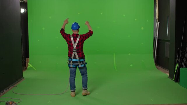 A male builder in professional ammunition and a construction helmet waving his hands and signaling to someone on a green background