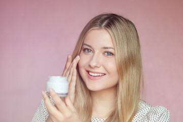 Smiling blonde woman applying moisturiser, holding face cream jar. Clear skin, blue eyes, beauty...