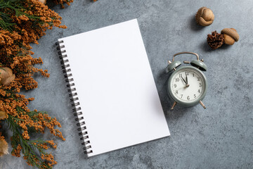 Notebook with blank pages and a vintage clock placed on a textured surface, accompanied by seasonal leaves and acorns for a cozy autumn atmosphere