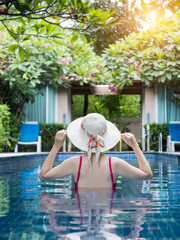 back view of woman in pink swimsuit with straw hat relaxing in swimming pool at hotel, enjoy summer holiday and vacation at tropical country, sunbathing wellness lifestyle concept.