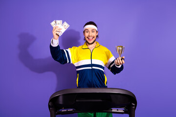 Excited athletic man celebrating victory holding money and trophy on treadmill against a vibrant...