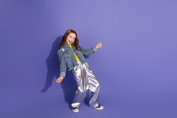 Fashionable young girl in trendy denim and metallic pants posing against a purple backdrop, showing...