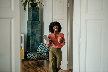 Smiling black woman walking through doorway holding laptop and coffee