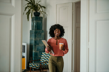 Smiling black businesswoman holding laptop and coffee walking through office doorway