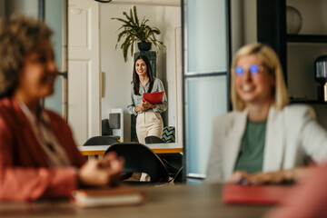Young businesswoman entering meeting room holding clipboard, colleagues waiting