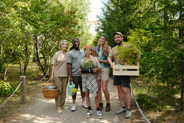 Group of diverse young adults and one teenager standing outdoors in garden holding potted plants and gardening tools, smiling and posing together for camera on sunny day