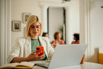 Businesswoman using smartphone and laptop in modern office with colleagues in background