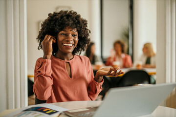 Smiling black businesswoman talking on phone in office with laptop and documents