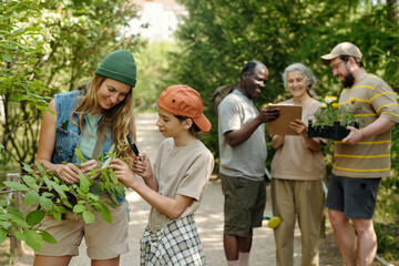 Caucasian young adult woman and Caucasian child examining leaves with magnifying glass outdoors, Black middle aged man holding clipboard, senior Caucasian woman and Caucasian man carrying plants