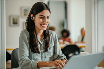 Smiling businesswoman wearing wireless earphones working on laptop in office