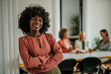 Confident businesswoman smiling with folded arms in modern office with coworkers meeting