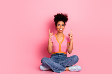 Young stylish woman on a pink background pointing upward with both hands while seated and wearing...