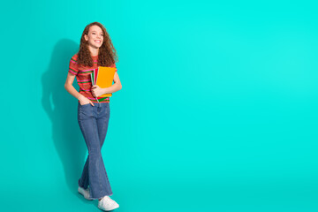 Cheerful young woman standing against a vibrant turquoise background holding notebooks and...