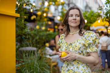 A woman in a yellow floral dress holds a lemon while standing in a lively outdoor area with string lights and greenery