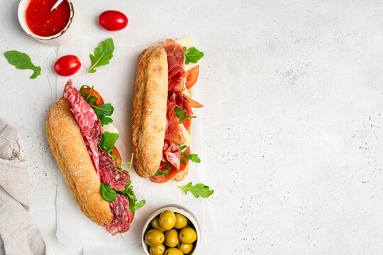 Assorted delicious baguette sandwiches filled with thinly sliced ham or salami and fresh green lettuce or arugula for breakfast arranged on white background, top view with copy space