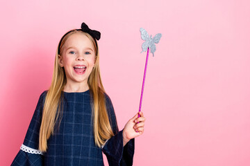 Cheerful young girl with blonde hair holding a butterfly wand in front of a pink background, wearing stylish blue dress