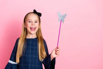 Fotobehang Cheerful young girl with blonde hair holding a butterfly wand in front of a pink background, wearing stylish blue dress © deagreez