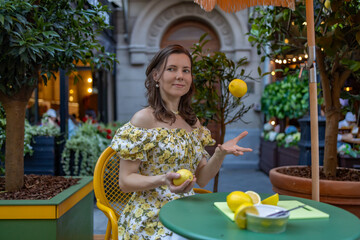 A woman in a floral dress juggles lemons while sitting at a table in a lively outdoor cafe surrounded by greenery and soft lighting