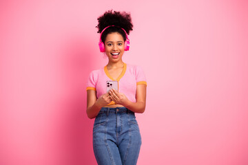 Stylish young woman wearing headphones holding smartphone against a pink background, joyful and...
