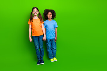 Two cheerful schoolgirls holding hands standing against a vivid green background, expressing...