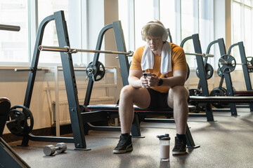 Sporty tired young caucasian man relax and sitting with mobile phone, listen music headphones on...