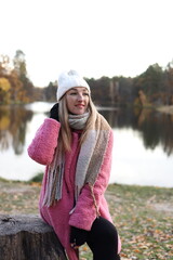 A woman sits on a log by a calm lake, wearing a pink sweater, hat, and scarf. The autumn colors of...