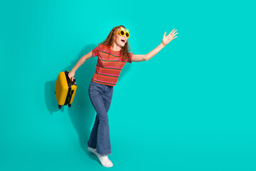 Stylish young woman with curly red hair holding a yellow suitcase, waving her hand, isolated on...