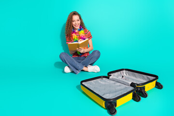 Happy young girl in casual wear preparing for travel with an empty suitcase sitting against a...