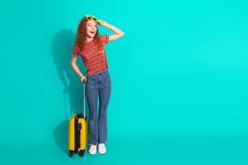Young cheerful woman with curly red hair and striped t-shirt posing with a suitcase against a...