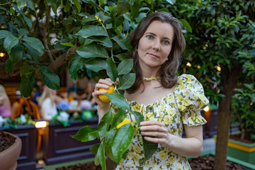 A woman in a yellow floral dress holds a lemon while standing in a vibrant garden with lush...