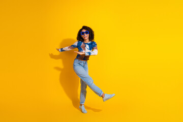 Cheerful young woman with curly hair wearing blue top and jeans expressing joy and posing against...